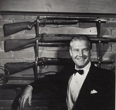 photo of George standing in front of three rifles 
displayed on the wall of his den.
