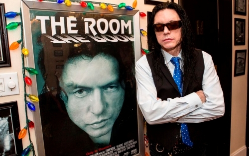 Tommy Wiseau standing by his poster for The Room
