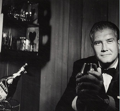photo of George, not smiling, just looking into 
the camera while holding a glass of brandy.